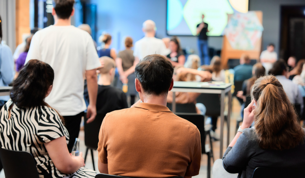 ki-einfuehrungsworkshop-guter-move KI Einführung für Wohlfahrtsverbände, Personen sitzen mit dem Rücken zum Betrachter und schauen auf eine Bühne auf der präsentiert wird, viele Menschen im Hintergrun verschwommen